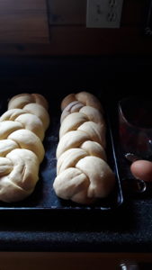 Two uncooked, braided egg bread loaves on pan, with fresh egg and measuring cup next to pan.